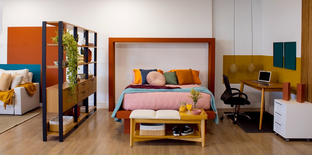 Quarto colorido integrado com uma sala é uma tendencia inverno 2022. Da esquerda para a direita temos a metade de um sofá aparecendo, uma estante marrom e preta dividindo o ambiente, uma cama de casal com colcha rosa e almofadas em tons de laranja, verde e azul, um banco aos pés da cama com um copo de suco em cima e, na lateral direita, uma escrivaninha com cadeira de escritório e gaveteiro.