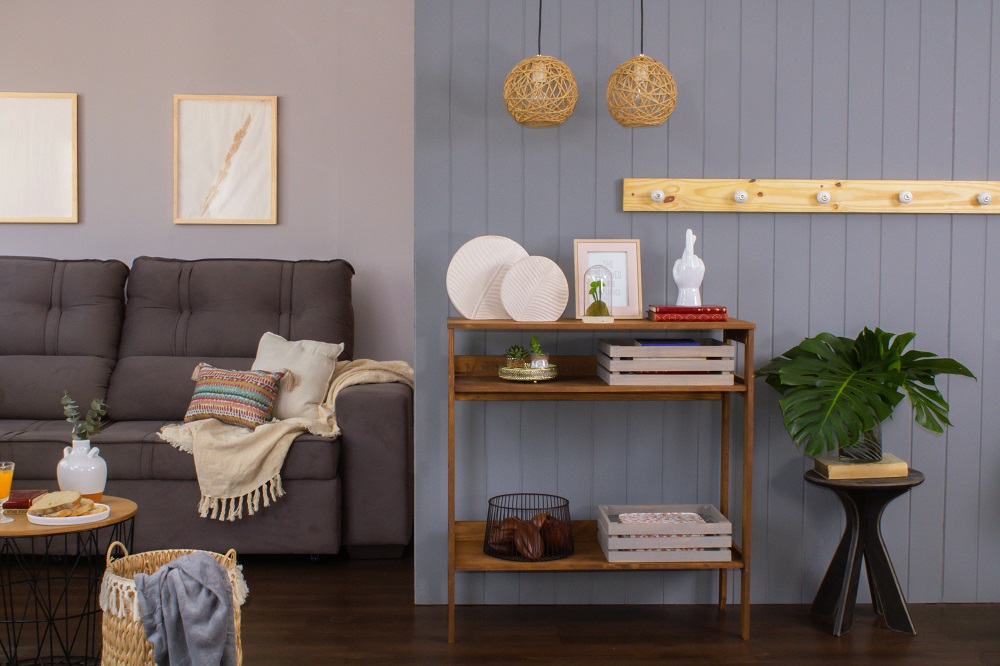 Sala de estar com metade de um sofá cinza aparecendo, assim como manta e almofadas em cima dele. Ao lado, temos um aparador com objetos decorativos, um banquinho servindo de apoio para um vaso de planta e um cesto de palha com uma manta saindo dele como tendencia inverno 2022