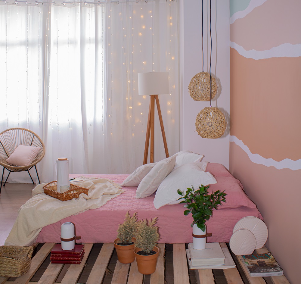 Quarto no estilo confy como tendencia inverno 2022 com cortina branca ao fundo, poltrona e abajur. Mais à frente, uma cama de casal com colcha rosa, almofadas brancas e uma bandeja de café da manhã apoiada.