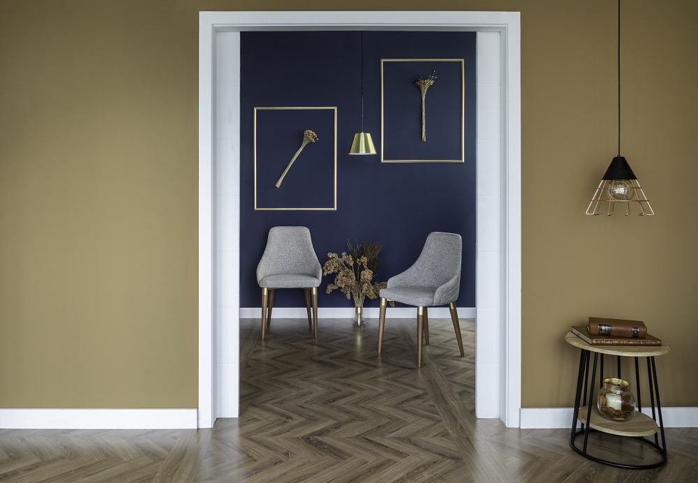 Imagem de uma sala com duas poltronas cinzas ao fundo. Na parede, que é azul, há duas molduras douradas com buquês de flores e um abajur centralizado. Mas à frente, a parede é mostarda. O dourado é uma tendencia inverno 2022