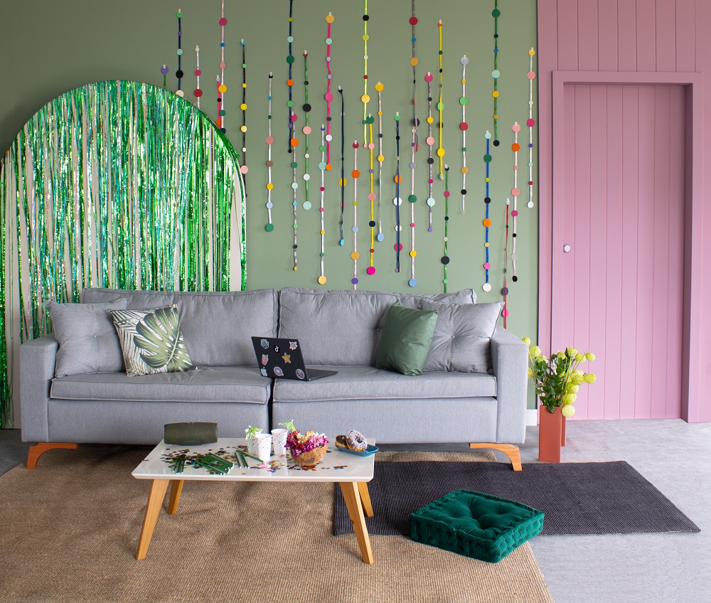 Sala de estar com parede ao fundo na cor verde e rosa. À frente um sofá cinza, mesinha de centro branca e objetos decorativos coloridos