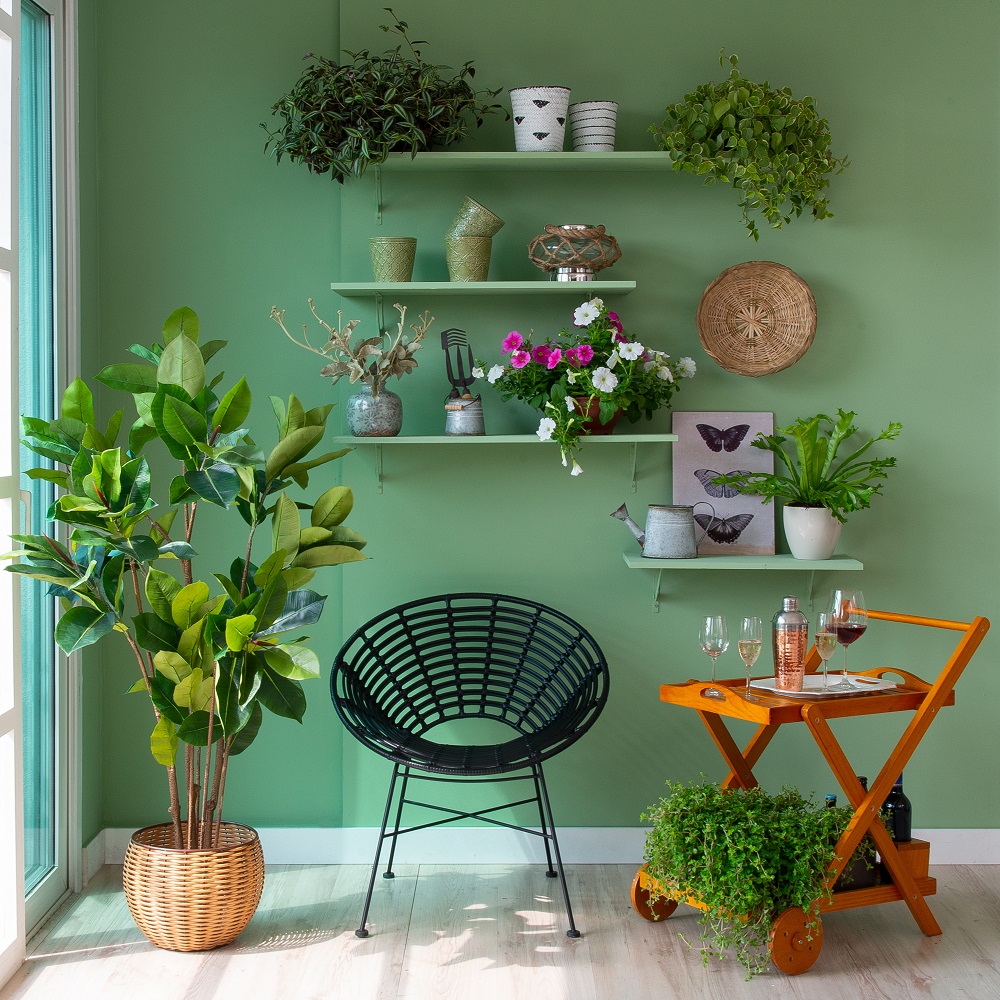 Cantinho da varanda com parede verde ao fundo, prateleiras com plantas, poltrona e carrinho bar.