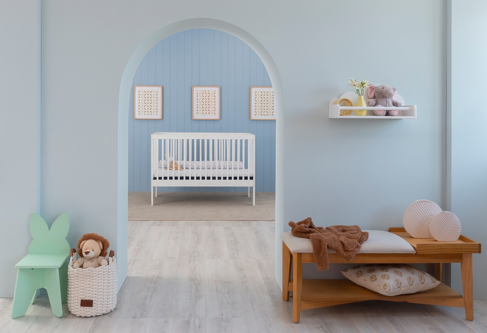Quarto infantil delicado com cores da primavera, um berço branco ao fundo, parede em dois tons de azul, um banquinho verde à frente e um banco 3 lugares em tom de madeira