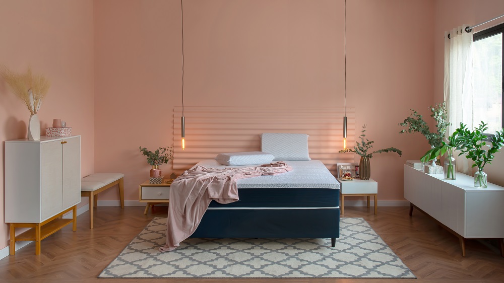 Quarto aconchegante em tons de rosa. Todas as paredes estão em rosa claro. Na lateral esquerda, há uma sapateira branca e um banco estofado. No meio, uma cama com colchão e coberta rosa acompanhada de duas mesinhas. Na lateral esquerda, um buffet branco com plantas no tampo.