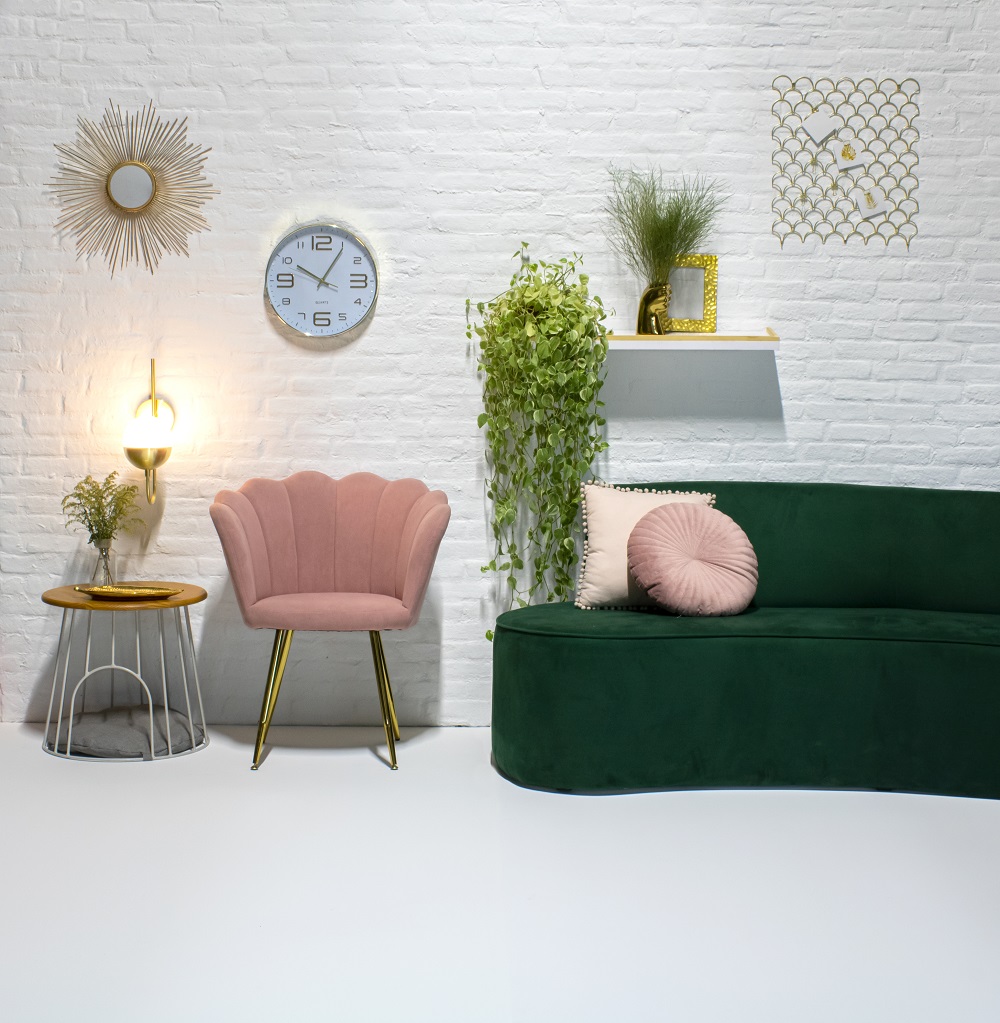 Fragmento de uma sala de estar com sofá verde e objetos decorativos rosados. A parede é branca com enfeites pendurados na cor dourada.