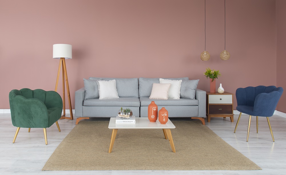 Sala de estar com parede rosa ao fundo, sofá cinza, poltronas na cor verde e azul. Tem um tapete bege no centro com uma mesinha branca sobre ele.