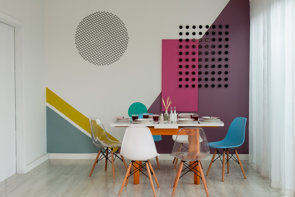 Sala de jantar colorida com cadeiras de diversas cores, mesa extensível branca e parede em rosa, roxo, branco, verde e amarelo.