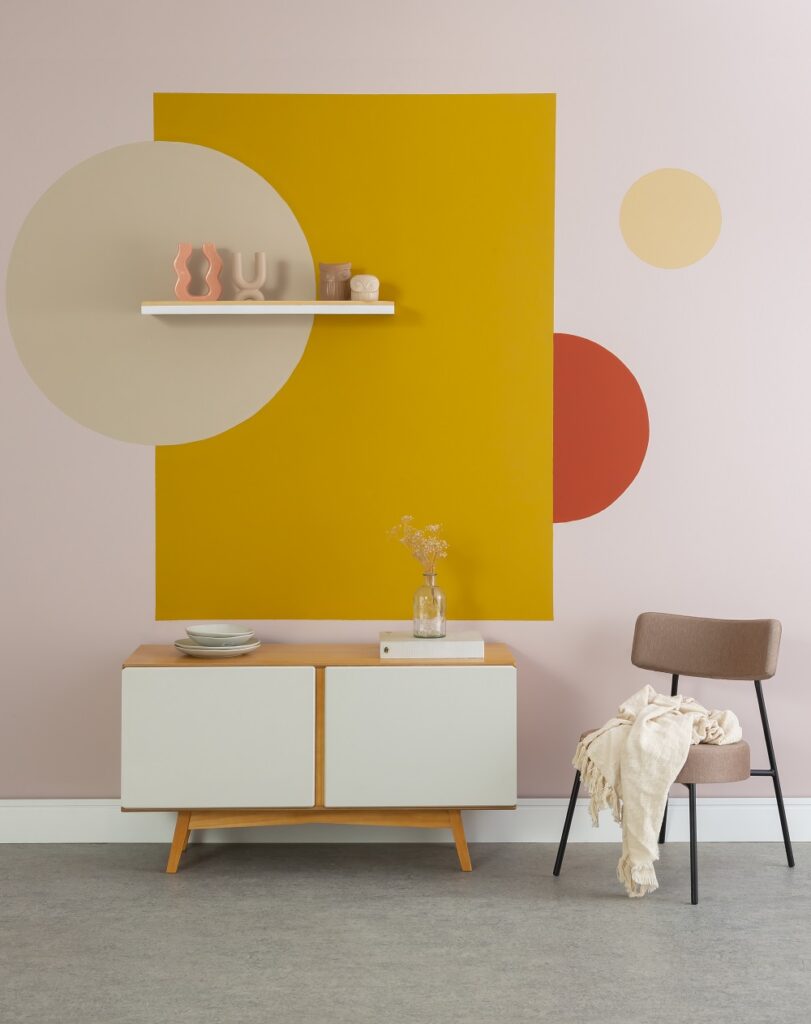 Fragmento de sala de jantar com parede rosa ao fundo e pinturas coloridas circulares e retangulares nas cores amarelo e laranja.