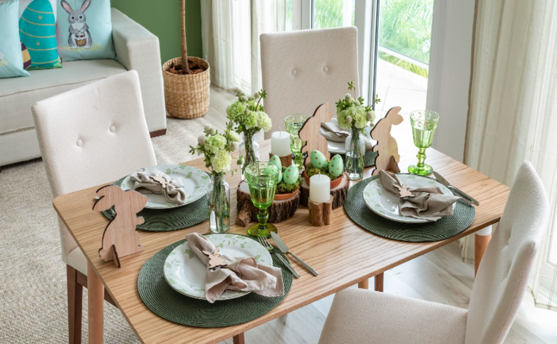mesa de páscoa com jogo americano, prato raso, vasos com plantas, adornos decorativos de páscoa, guardanapo de tecido e talheres