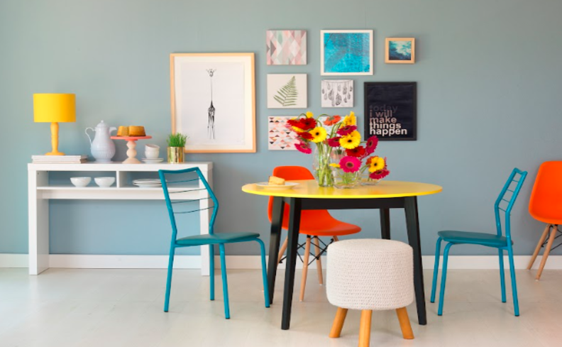 tons de azul pastel nas cadeiras em um ambiente de sala de jantar, além de mesa de madeira, puff e um aparador, além de quadros decorativos.