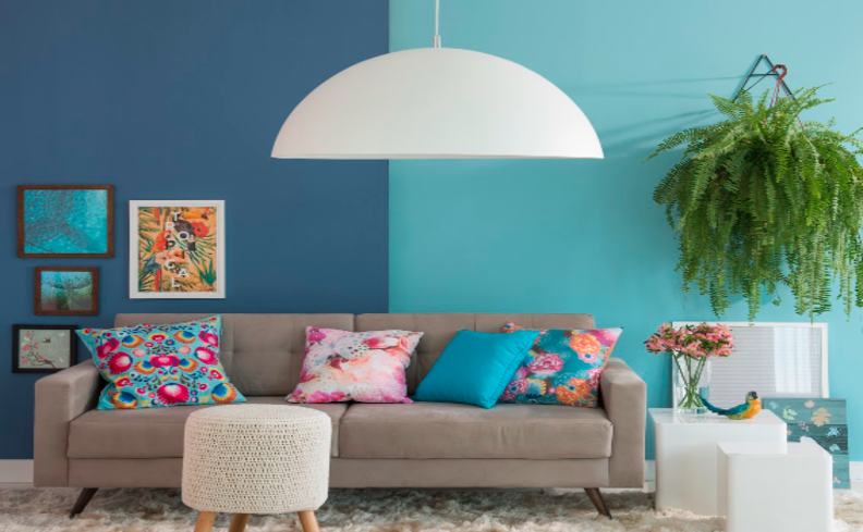 dois tons de azul para parede em uma sala de estar com sofá cinza, almofadas coloridas, puff e quadros decorativos.