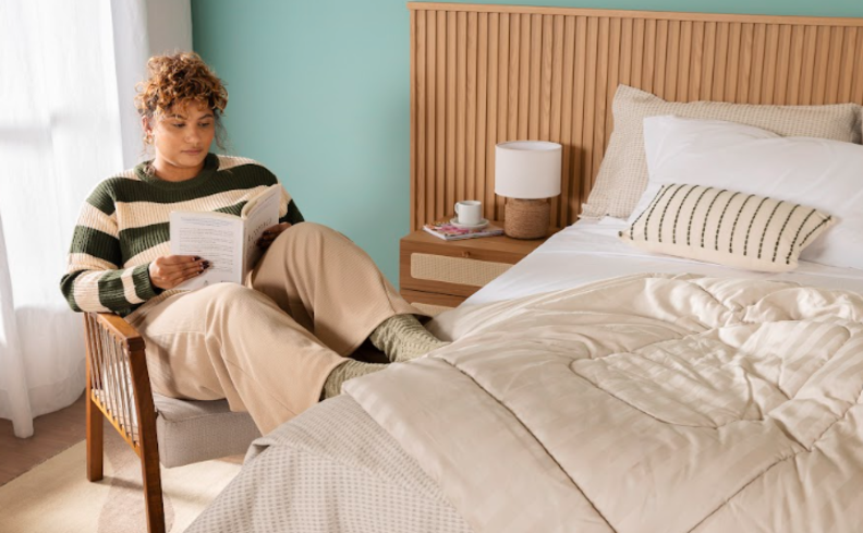 tipos de cortina para quarto com mulher sentada em uma poltrona lendo, além de mesa de cabeceira e cama posta completa.