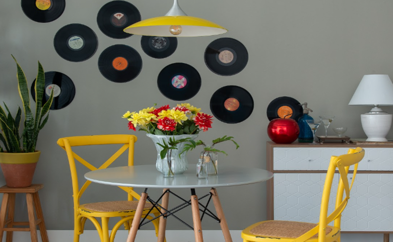 decoração com disco de vinil na parede da sala de estar, além de mesa redonda, cadeiras amarelas, vasos com plantas e um buffet com um abajur.