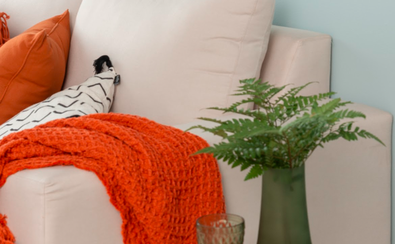 laranja na decoração em um sofá off white, com manta para sofá e almofada laranja, além de vaso com planta.