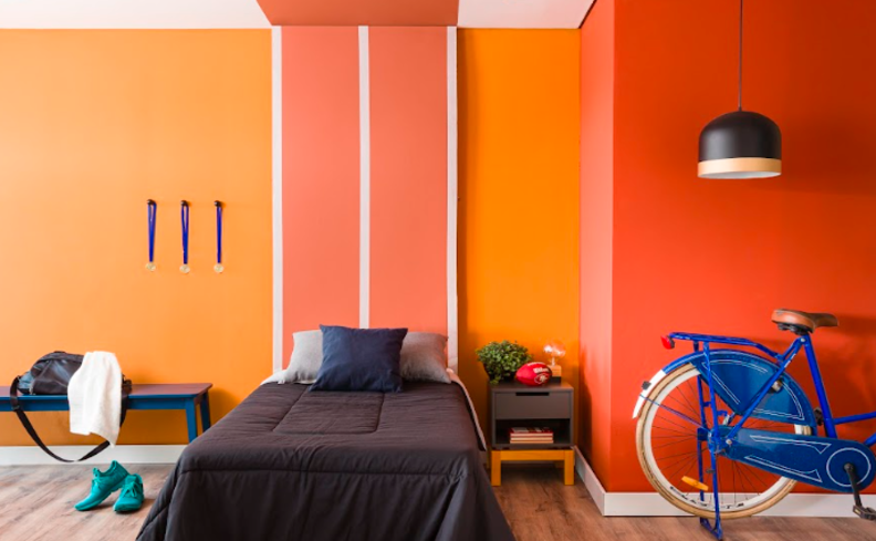 tons de laranja na parede do quarto, com cama, bicicleta na decoração, lustre pendendente e um banco de apoio.