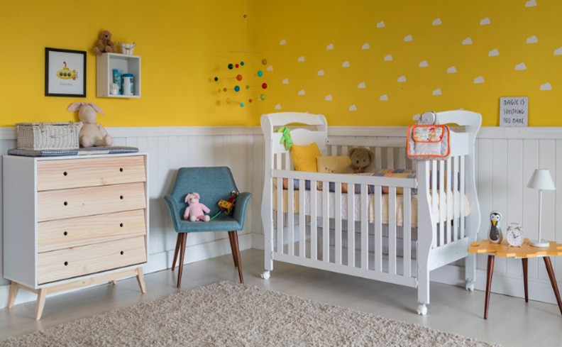 cômoda trocador de bebê, além de berço no quarto infantil, poltrona, mesa de apoio, parede amarela e branca e itens decorativos para deixar o espaço bonito e aconchegante.