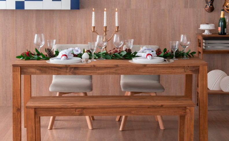 mesa posta de natal completa com mesa de madeira, banco e duas cadeiras brancas, além de louças, talheres, velas e taças.