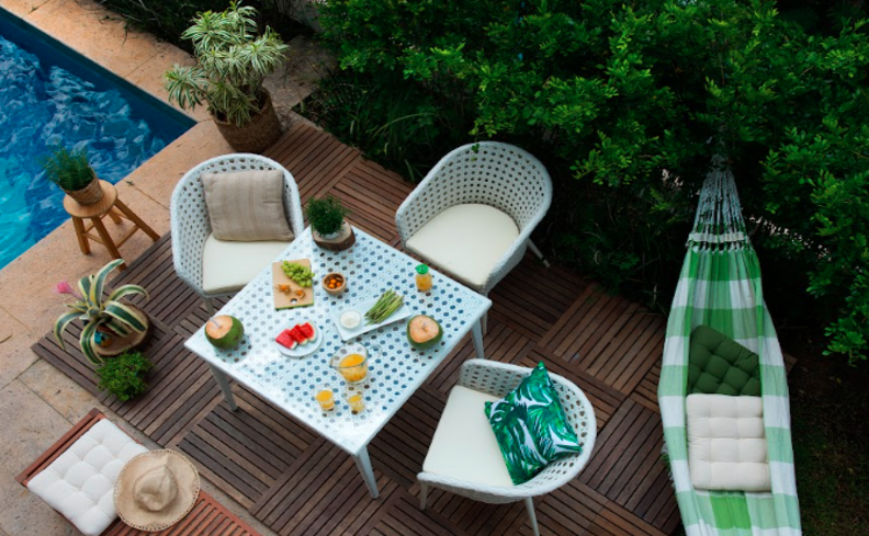 decoração varanda pequena com mesa quadrada, três cadeiras, rede, almofadas decorativas e diversas plantas.