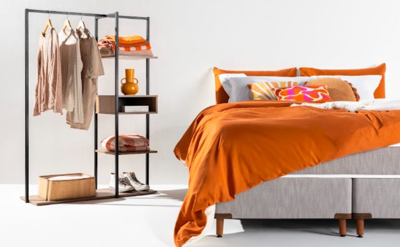 quarto de casal com cama posta com tons de laranja, arara de roupas, e diversos travesseiros e almofadas na cama posta.