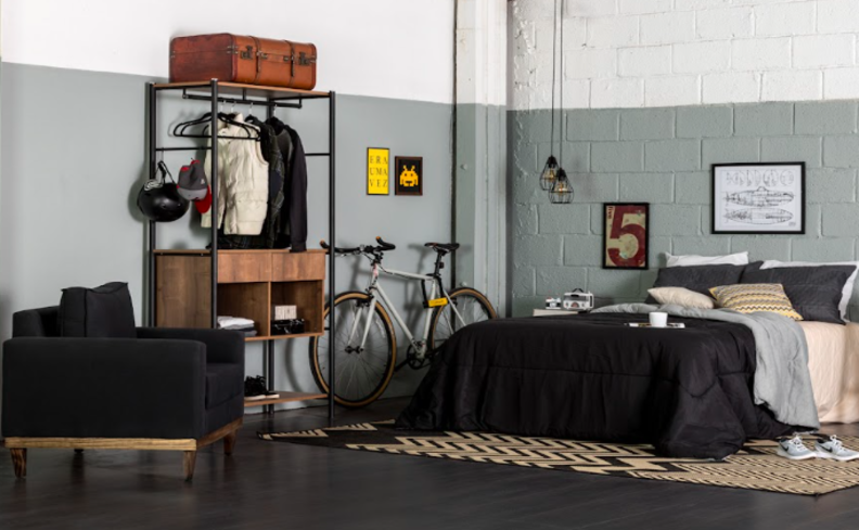 quarto estilo industrial com arara de roupas com base de metal preta, cama com roupa de cama preta e cinza, parede cinza e branca, além de pendentes e quadros decorativos para complementar.