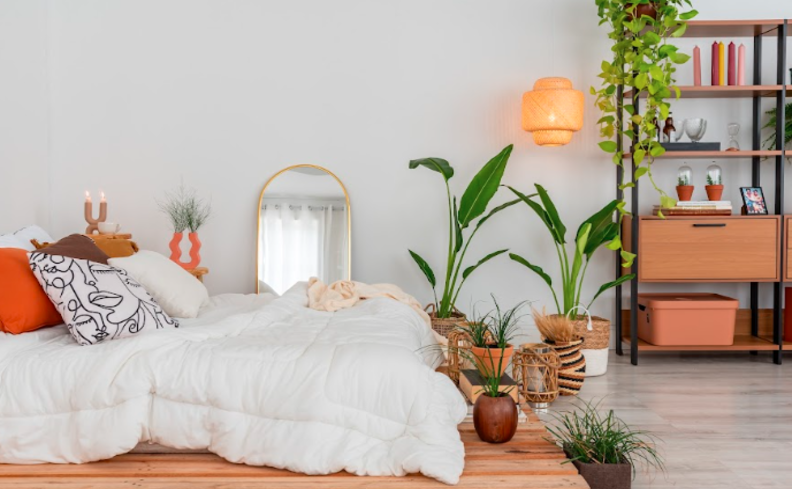 luminária de teto para quarto em tons claros em madeira, branco e alguns toques vibrantes em detalhes como almofadas e outros adornos decorativos, além de uma estante e plantas.