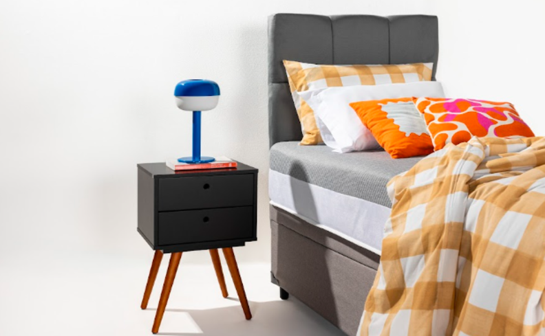 luminária para quarto em um tom de azul, mesa de cabeceira preta, cama de solteiro com cabeceira cinza e roupas de cama em tons de branco e laranja.