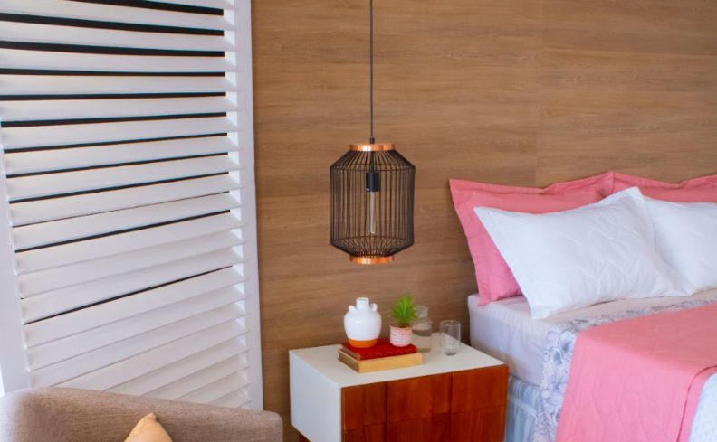 ambiente com luminária pendente para quarto, mesa de cabeceira, cama de casal com roupas de cama em tons de branco e rosa, além de uma poltrona e almofada.