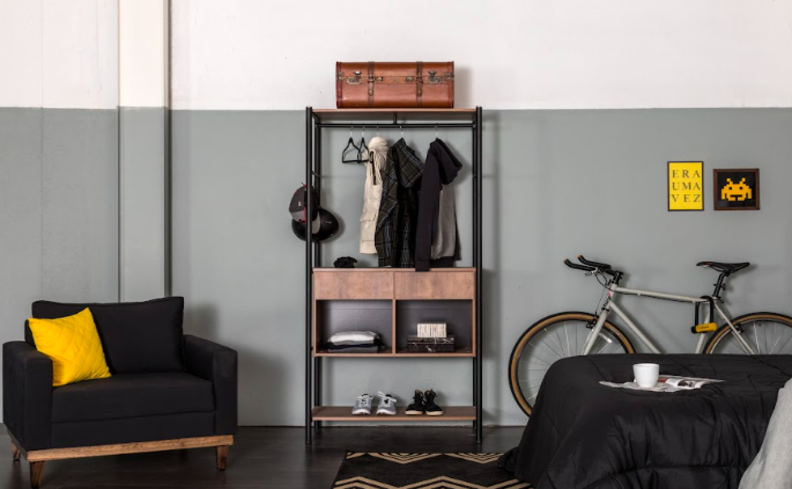 arara de roupas com rodinhas em uma estrutura de metal preto, com nichos, além de poltrona preta, cama de solteiro e uma bicicleta dentro do ambiente.