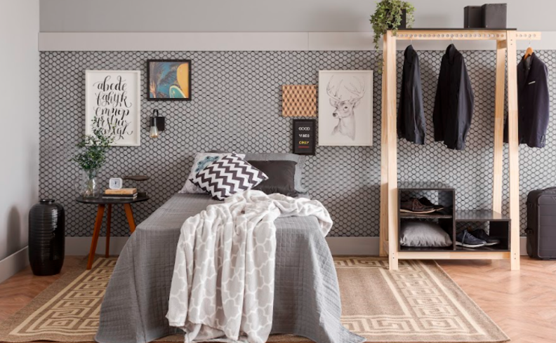 arara de roupas de madeira em um quarto com cama de solteiro com edredom cinza, almofadas e travesseiros, quadros decorativos, tapete e mesa de cabeceira.