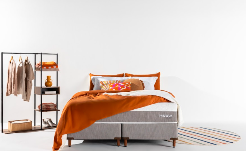 modelo de araras para roupas em um quarto com cama e roupa de cama em tons de laranja, além de uma arara em ferro preto com diversas peças organizadas no móvel.