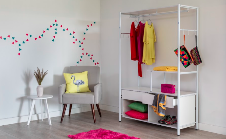 quarto com arara de roupas em metal na cor branca, além de nichos para conseguir organizar as peças de roupas. Ambiente também com poltrona e mesa de apoio.
