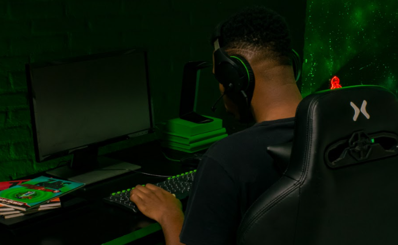 homem usando um headfone sentado em uma cadeira ergonômica gamer para jogar com conforto em uma mesa com computador e LED verde ao fundo.