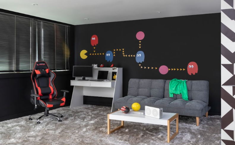 sala de estar com cadeira gamer preta e vermelha, mesa para computador, sofá cinza, mesa de centro branca e uma parede preta com desenhos do jogo pac-man para compor um setup gamer cheio de personalidade.