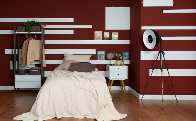 cores vibrantes que combinam entre si, com parede vinho com branco, luminária de chão preta, cama com edredom branco e diversas almofadas, arara de roupas, mesa de cabeceira branca de madeira e itens decorativos para complementar.