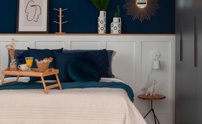 decoração apartamento pequeno simples no quarto com cama, roupa de cama azul e branca, prateleira com espelho, diversos quadros e adornos decorativos, além de uma mesa de cabeceira com abajur.