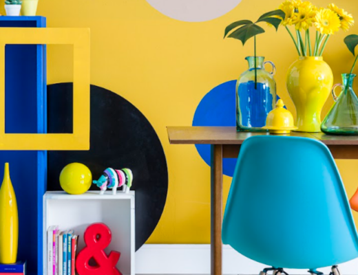 decoração maximalista na sala de jantar com mesa de madeira, parede amarela com desenhos de círculos preto, branco e azul, estante azul e amarela, cadeira azul e diversos itens decorativos.