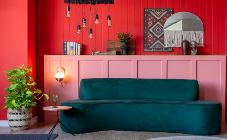 decoração maximalista na sala de estar com sofá verde, parede vermelha e rosa, diversos quadros decorativos, lustre pendente grande, vaso com planta e outros itens de decoração.
