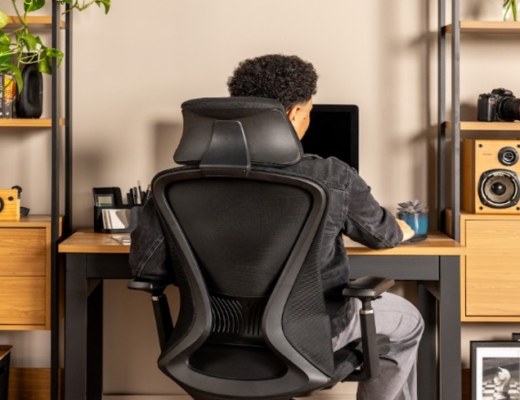 mesa com regulagem de altura com um homem sentado em uma cadeira de escritório, em um ambiente com mesa, estantes e diversos itens decorativos.