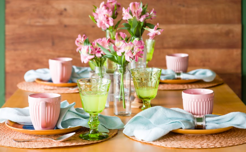 mesa posta dia das mães com xícaras rosas, jogo americano, talheres, guardanapo de tecido azul e um arranjo de vaso com flores no centro da mesa.