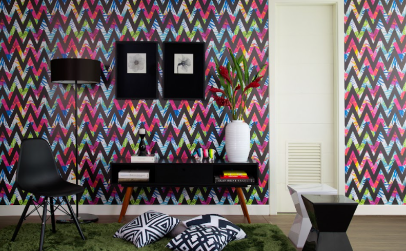 o que é maximalismo em uma sala de estar com parede com cores vibrantes e estampa geométrica, móveis pretos e brancos e um tapete verde, além de diversos itens de decoração.