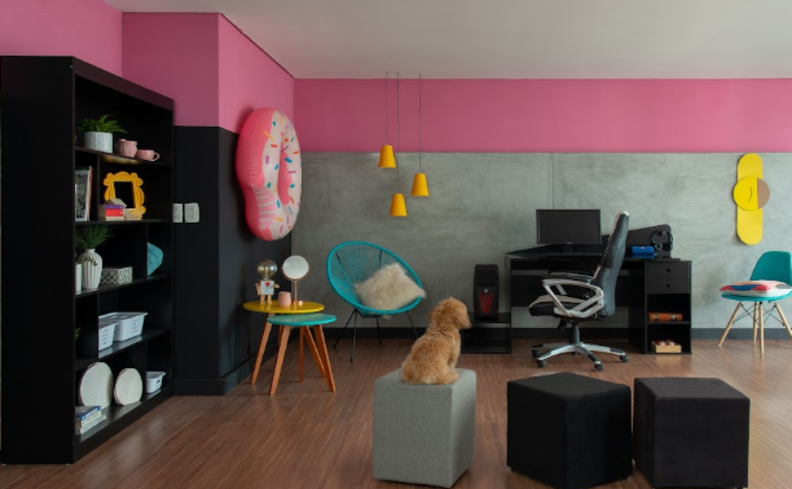 quarto maximalista com parede rosa e cinza, móveis pretos, uma boia como item decorativo, mesa lateral, escrivaninha, cadeira para escritório, poltrona e uma estante com diversos itens decorativos.