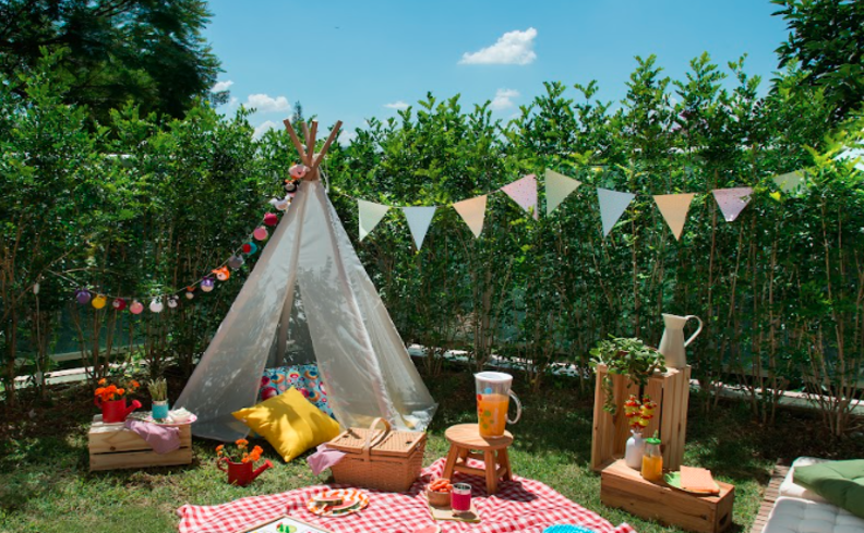 decoração de mesa para festa junina em casa no jardim com diversas bandeirinhas, tapete para fazer um piquenique, almofadas, uma mesa de apoio e diversas comidas típicas da época.