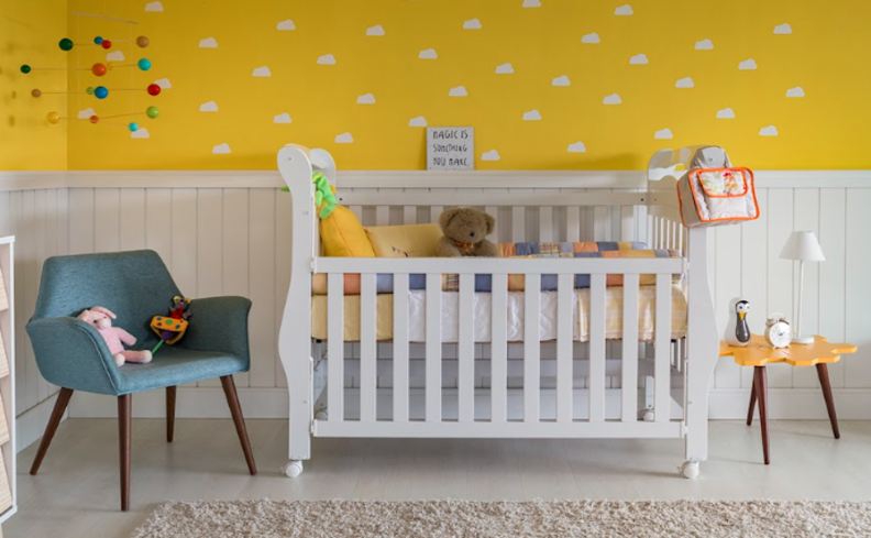 decoração para quarto de bebê menina e menino com meia parede amarela e branca, berço branco, poltrona azul, mesa lateral amarela e diversos ursinhos de pelúcia.