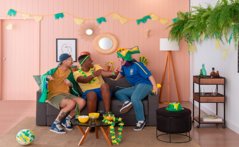 decoração brasil com três amigos sentados no sofá comemorando os jogos da seleção ne sala de estar com sofá, estante pequena, luminária de chão e uma decoração de copa do mundo