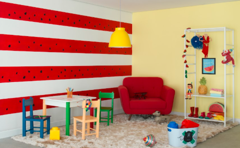 cantinho de estudos infantil com mesa, duas cadeiras, poltrona vermelha, parede com listras vermelhas, estante branca e diversos brinquedos infantis.