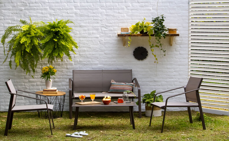 cantinho do churrasco pequeno e simples na área externa da casa com sofá, duas poltronas, prateleira e diversos vasos com plantas.