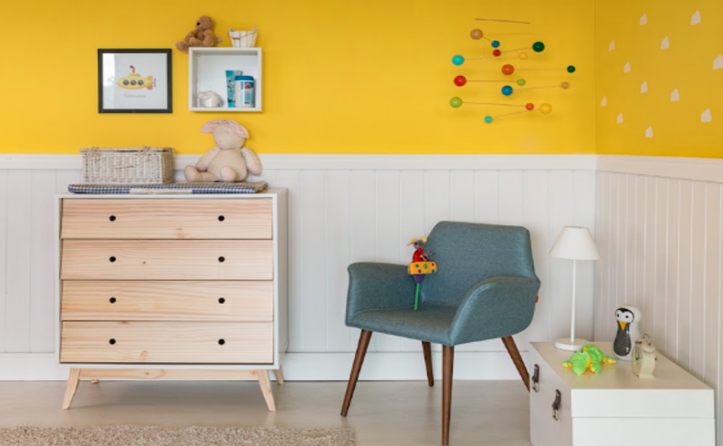 decoração quarto infantil simples com cômoda de madeira, poltrona azul, uma parede metade amarela e a outra metade branca e diversos itens decorativos.
