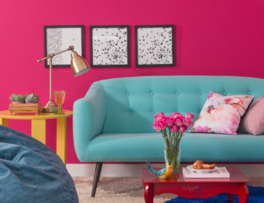 móveis coloridos para ter uma decoração colorida, com sofá azul claro, pufe azul escuro, parede pink, mesa lateral rosa e amarelo, mesa de centro vermelha e quadros decorativos, almofadas e flores.