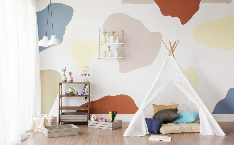quarto de brinquedos com cabana no chão, parede com diversos desenhos coloridos, mesa de apoio e várias almofadas.