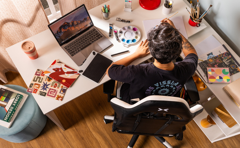 decoração home office quarto com escrivaninha branca, mulher sentada em uma cadeira gamer trabalhando, diversos itens de trabalho e escritório sobre a mesa.